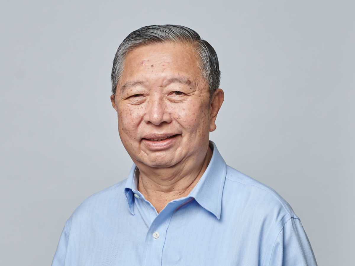 Mr Tan Soo Nan in a light blue shirt smiling with hands loosely clasped against a plain grey background